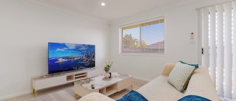A modern living room with a flat-screen TV, a sofa, a coffee table, and a window with blinds.