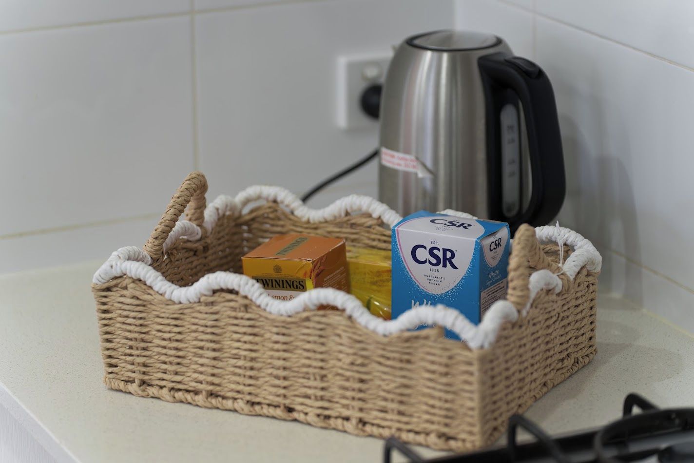 Un panier en osier contenant une boîte de thé Twinings, une bouteille de miel et une boîte de café CSR, posés sur un plan de travail de cuisine.