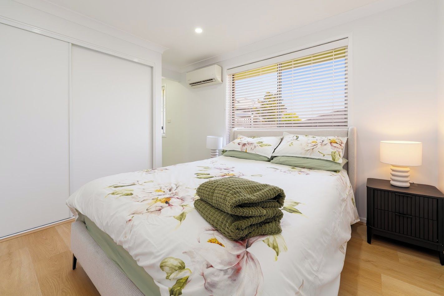 A bedroom with a bed, a bedside lamp, a window with blinds, and an air conditioning unit.