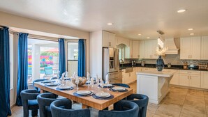 A modern kitchen with white cabinets, a central island, and a dining area with blue chairs and a wooden table set for six. - Peoria Resort! Spacious 7BR, Heated Pool, Theater, Hot Tub, Ping Pong & More! (Peoria)