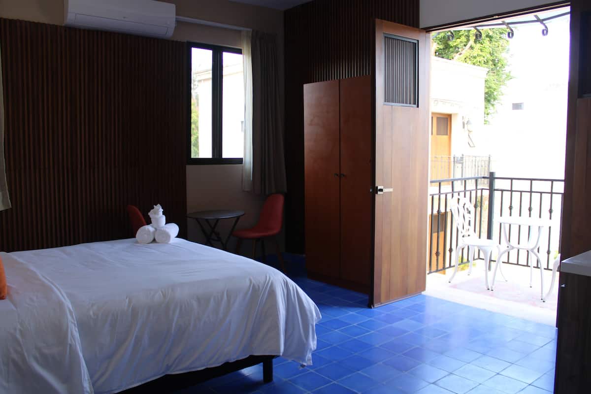Habitación de hotel con cama, una mesita, una silla roja, un armario y un balcón con barandilla.