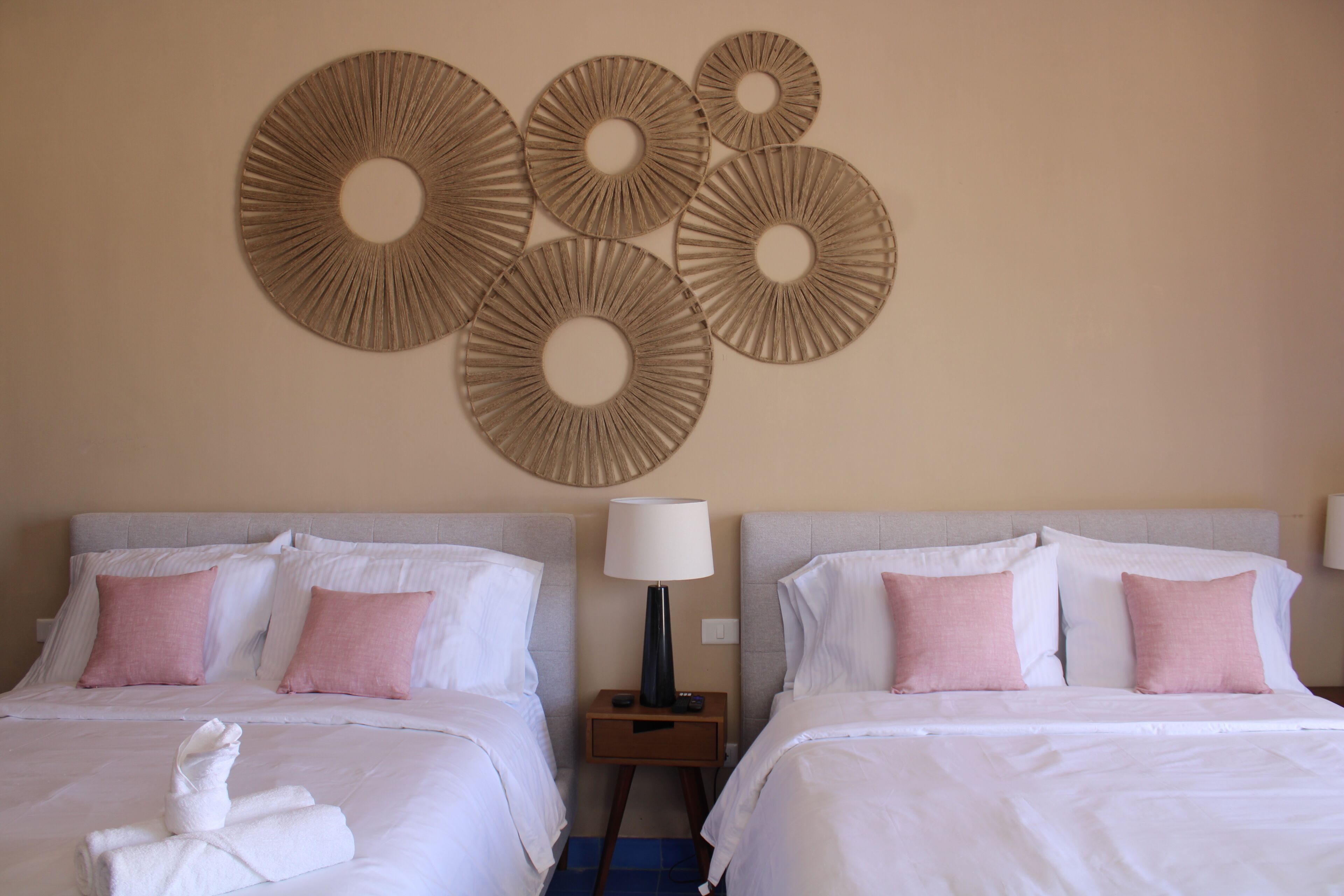 A hotel room with two beds, each with white bedding and pink pillows, a wall-mounted decorative piece, and a nightstand with a lamp.