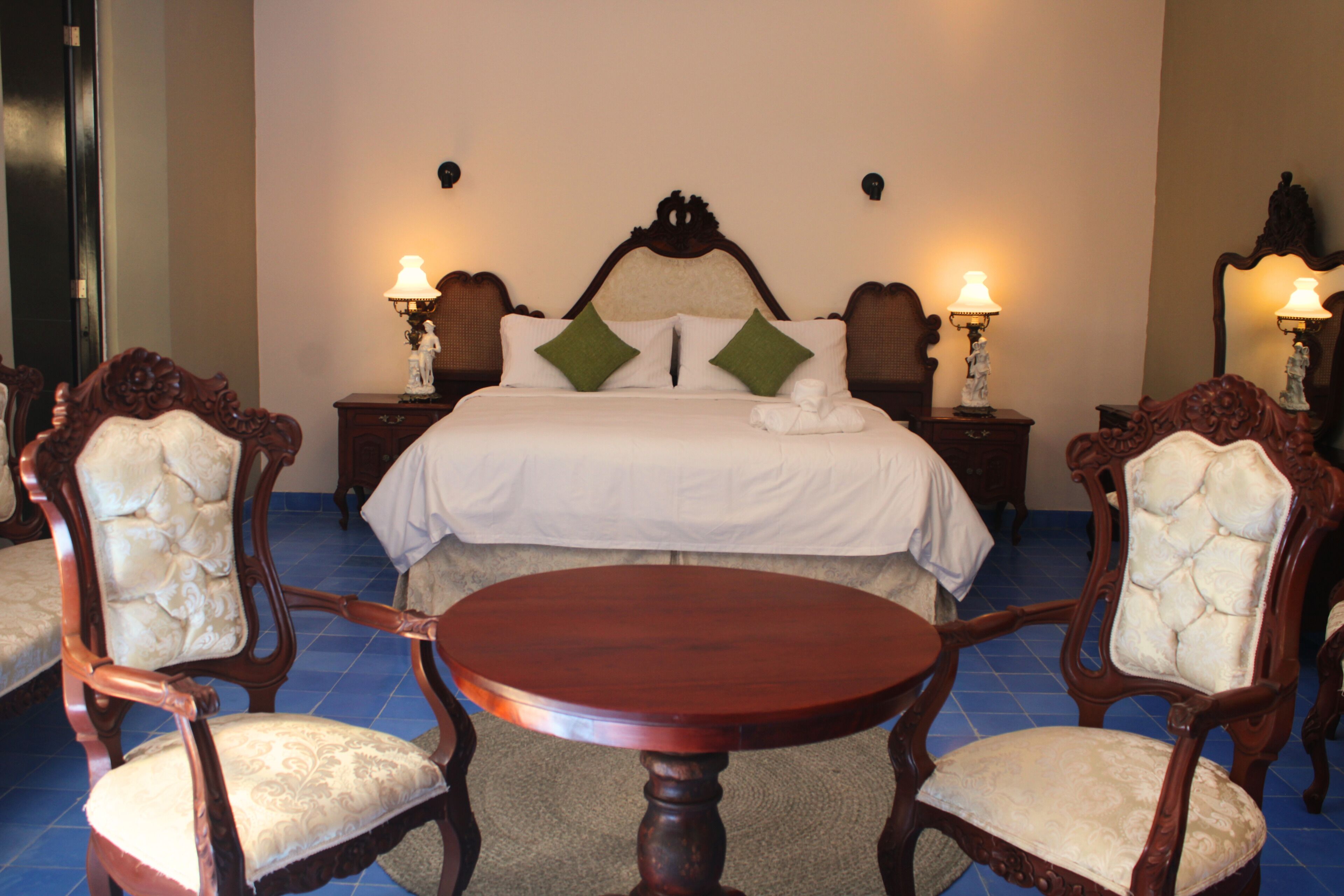 Quarto de hotel com uma cama grande, duas mesas de cabeceira com abajures, uma mesa de madeira redonda e duas cadeiras estofadas.