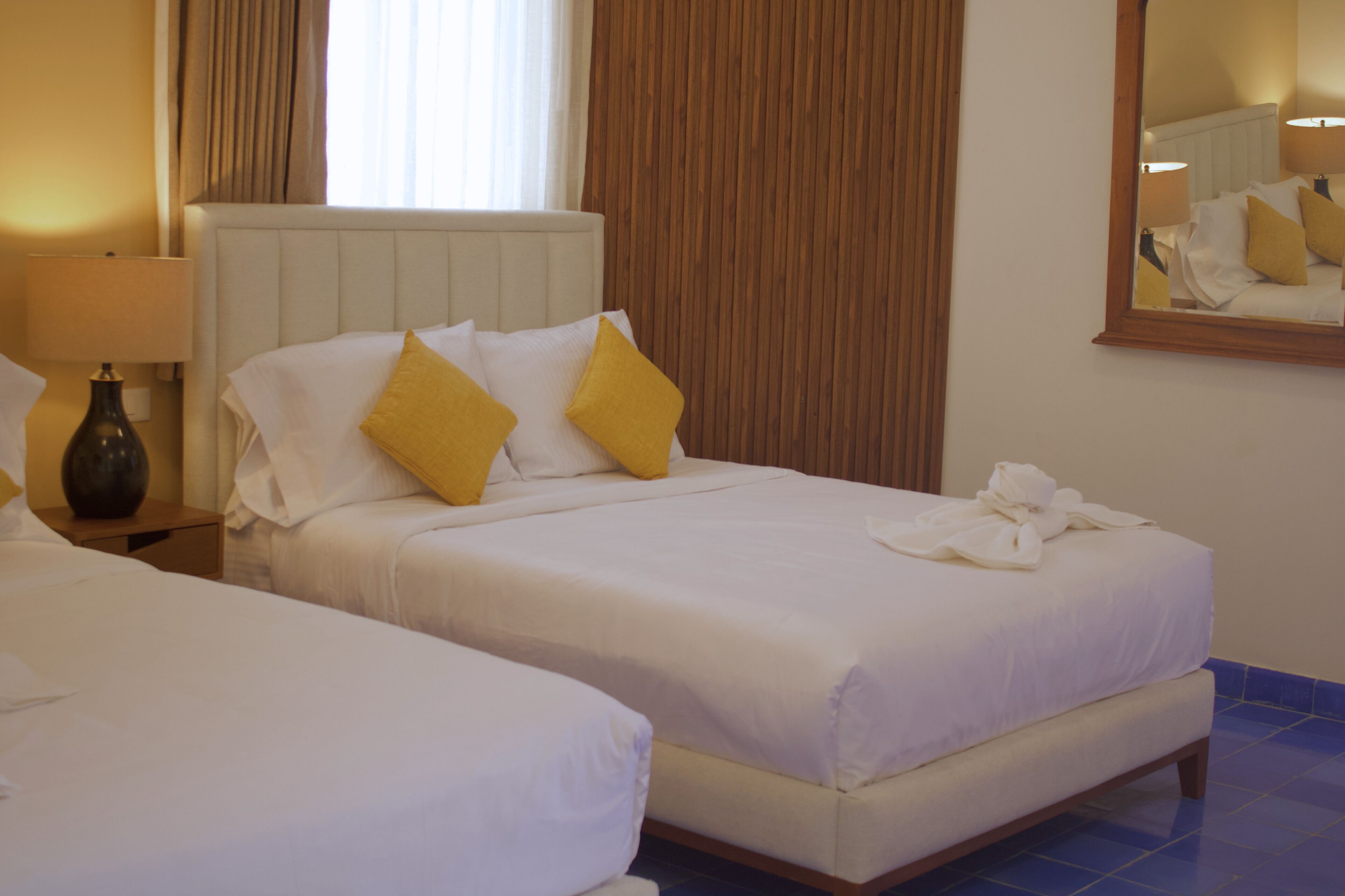 Two single beds with white linens and yellow pillows, a bedside lamp, and a mirror on the wall.