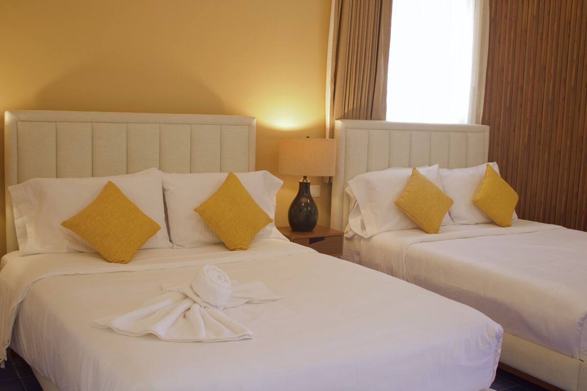 Dos camas individuales con ropa de cama blanca y almohadas amarillas, una mesita de noche con lámpara y una ventana con cortinas.