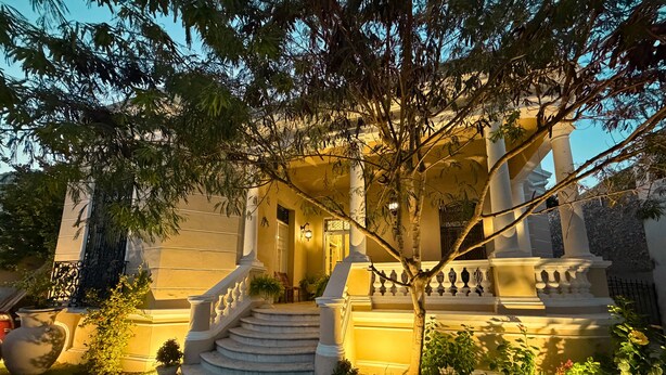 A grand entrance with a staircase leading to a porch, surrounded by lush greenery and well-maintained lawns. - Casa Ku'x (Merida)