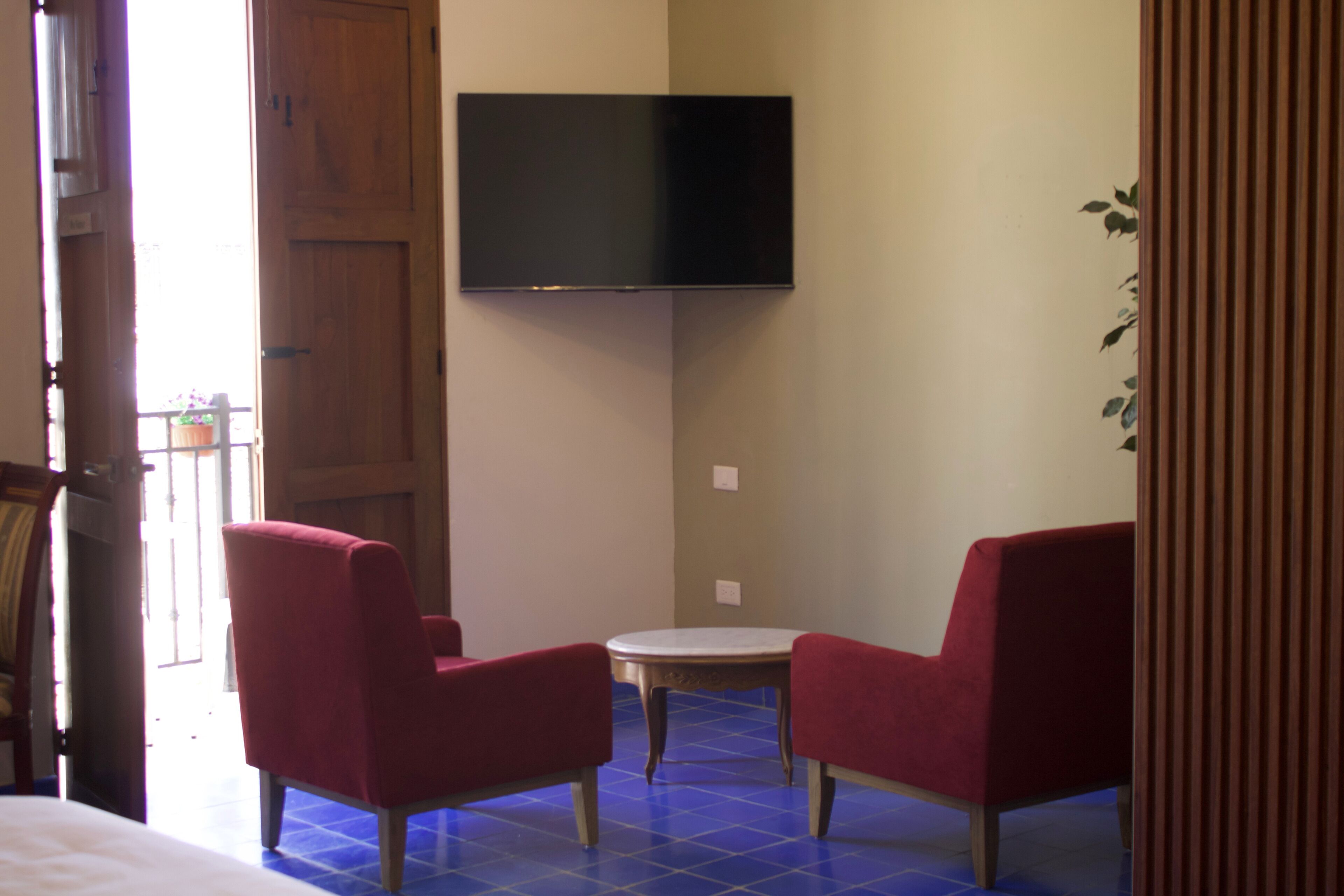 Une pièce comprenant un lit, un téléviseur fixé au mur, deux chaises rouges, une petite table ronde et un sol recouvert de carreaux bleus.