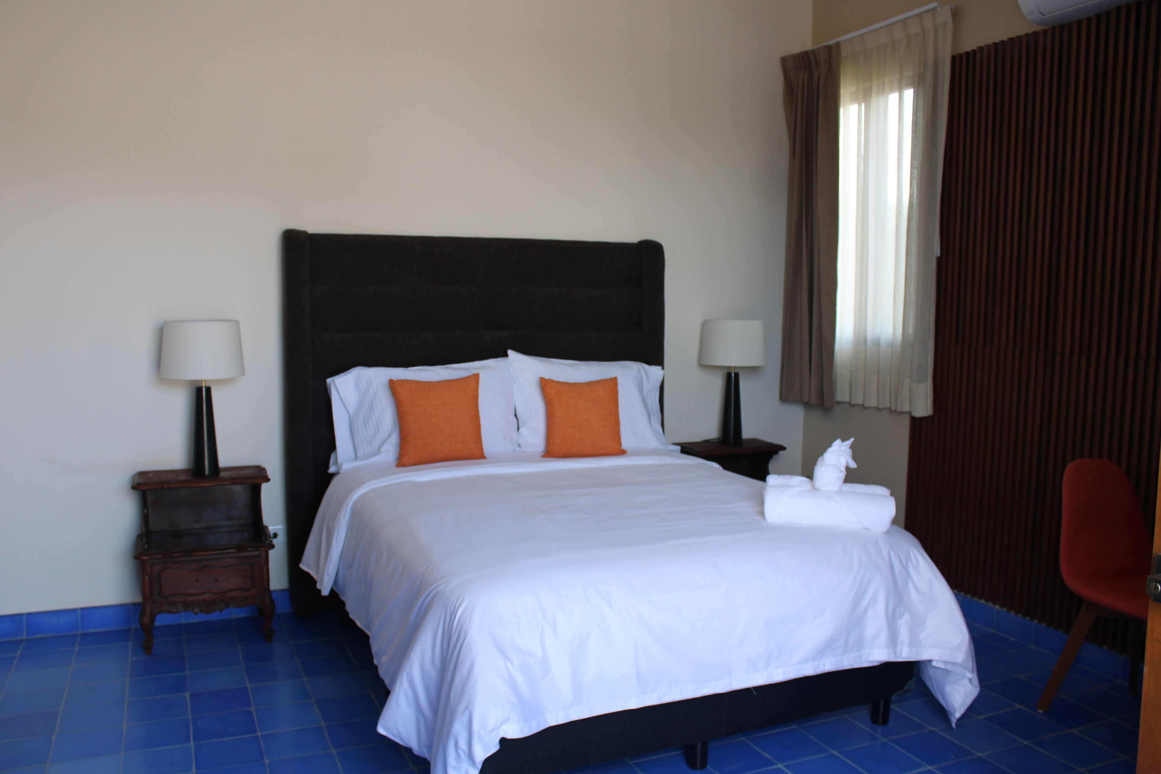 Una cama con ropa de cama blanca y almohadas naranjas, dos mesitas de noche con lámparas, una silla roja y una ventana con cortinas.