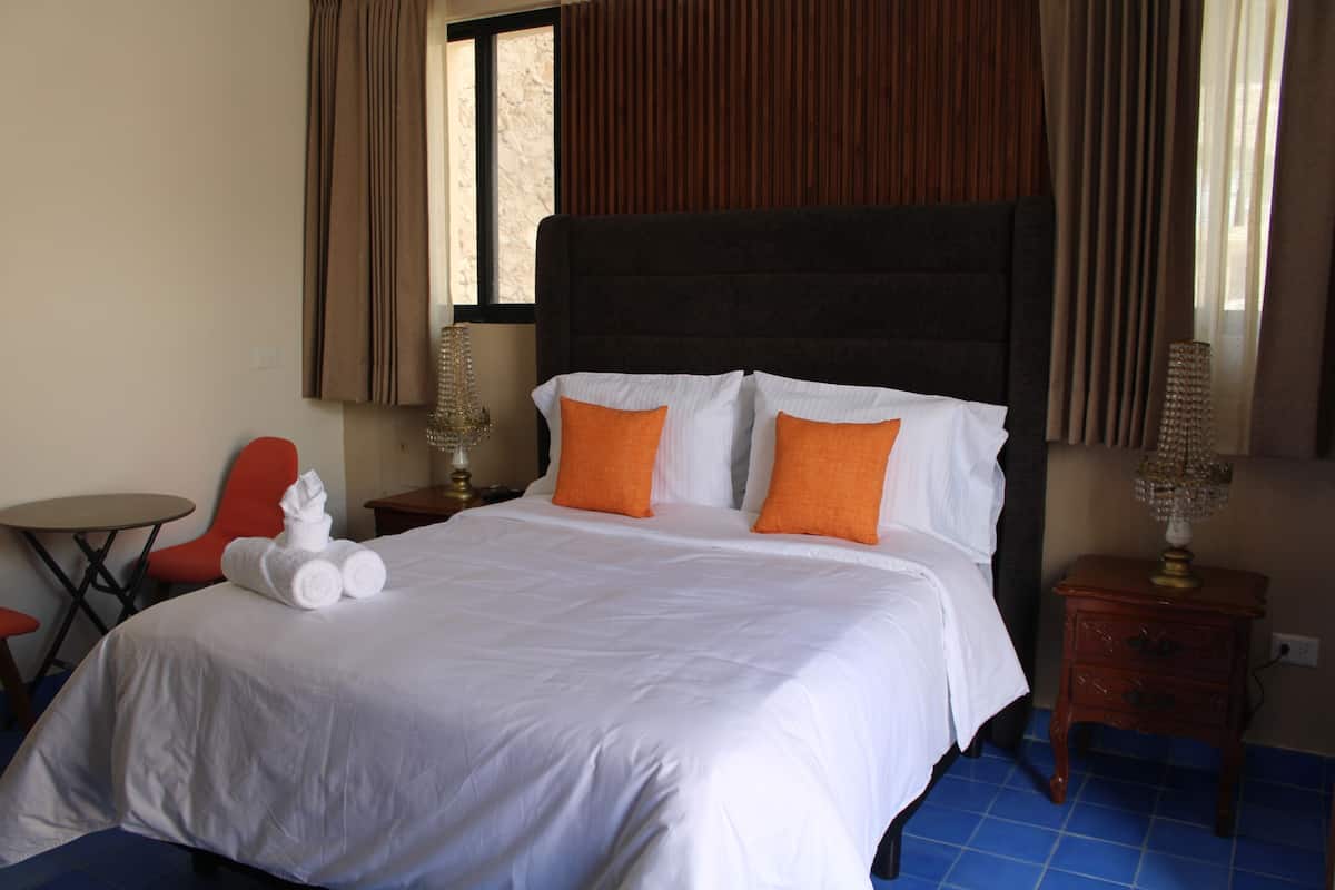 Habitación de hotel con una cama grande, dos almohadas, una mesita de noche con lámpara, una silla y una pequeña mesa.