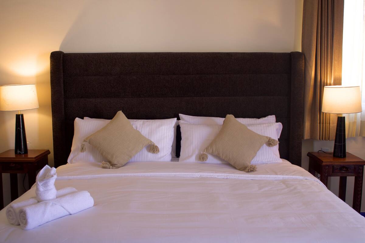 Una cama bien hecha con sábanas blancas y dos almohadas beige, flanqueada por dos mesitas de noche con lámparas.