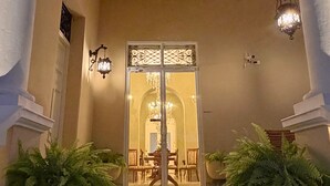 A grand entrance with a glass door, flanked by columns and potted ferns, leading to an interior space with a chandelier and a dining area. - Casa Ku'x (Merida)