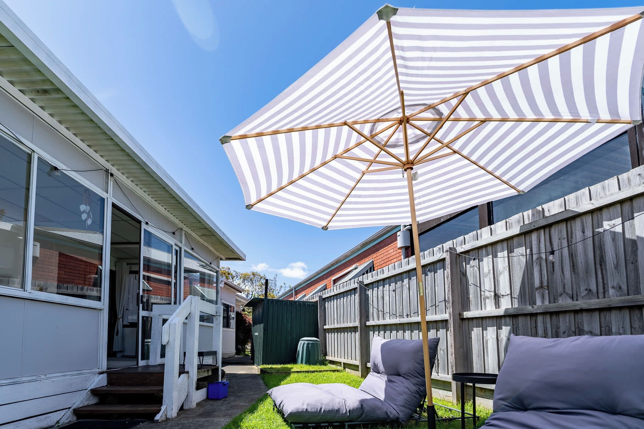 A backyard with a wooden fence, a striped umbrella, and a cushioned seating area.