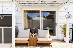 An outdoor seating area with a wooden table and two cushioned chairs, a grill, and a potted plant. - Classic 70's Luxury Shack | Pet Friendly (Ocean Grove)