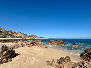 A coastal beach with rocky formations, clear blue sky, and a distant hill with buildings. - Modern Desert Beach Retreat, Rincon del Afan (Palmilla)