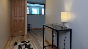 Un pasillo con puerta de madera, una mesa con lámpara, alfombra blanco y negro, y una ventana con persianas.