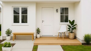 A white house with a front door, two windows, a bench, and potted plants.