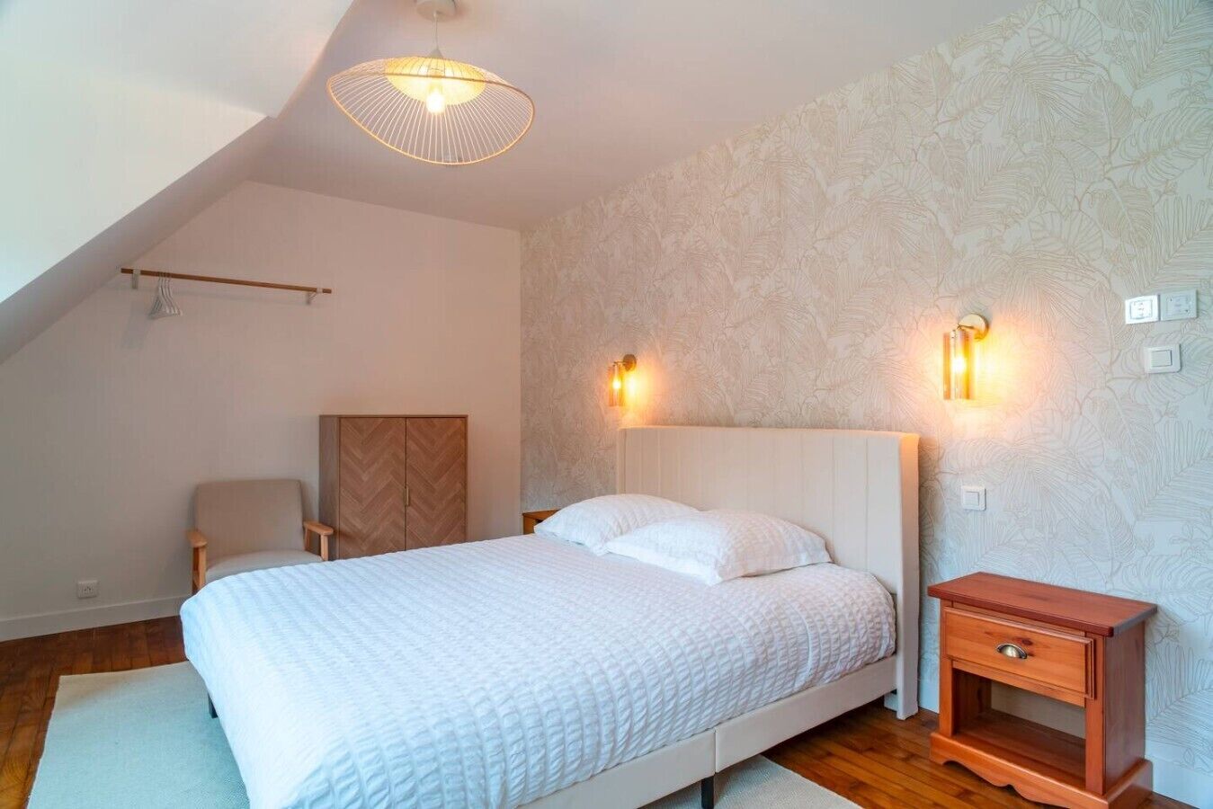 Ein Schlafzimmer mit Bett, Nachttisch aus Holz, Stuhl, Kleiderschrank und Deckenleuchte.