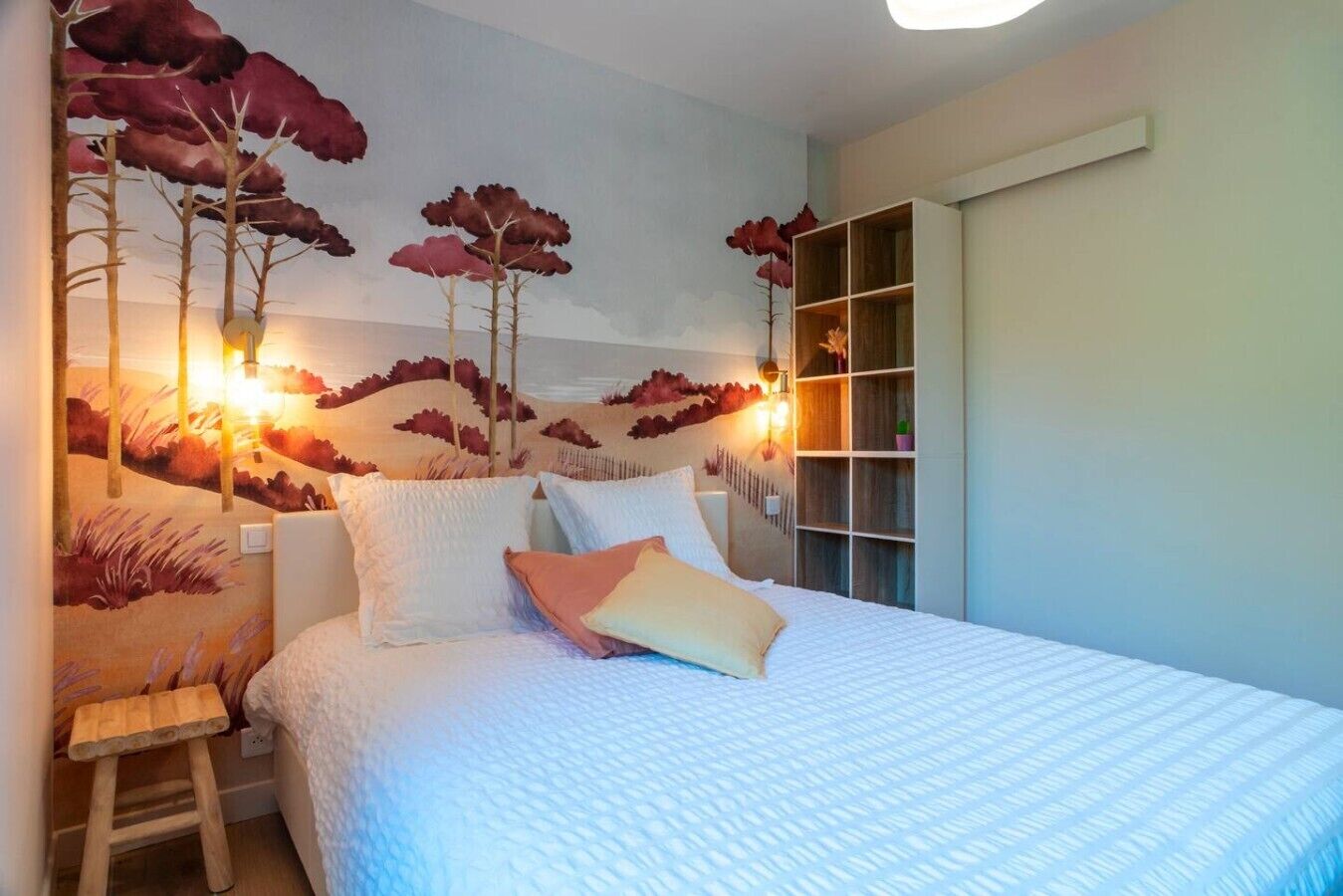 Un dormitorio con cama, almohadas, una mesita de noche, un taburete de madera y una pared decorada con un mural de temática natural.