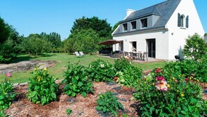 Una casa blanca con techo oscuro, un porche cubierto y un jardín con diversas plantas y flores.