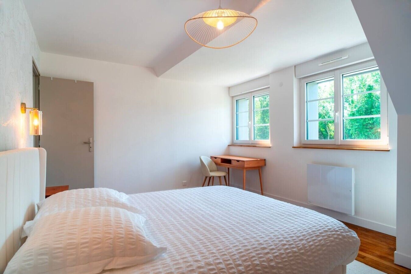A bedroom with a bed, a desk, and a window with a view of greenery.
