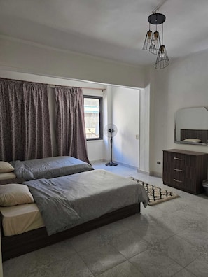 A bedroom with two beds, a standing fan, a window with curtains, a bedside table, and a dresser. - 2-bedroom apartment in marvelous Heliopolis with AC , WIFI and all needs . (Heliopolis)