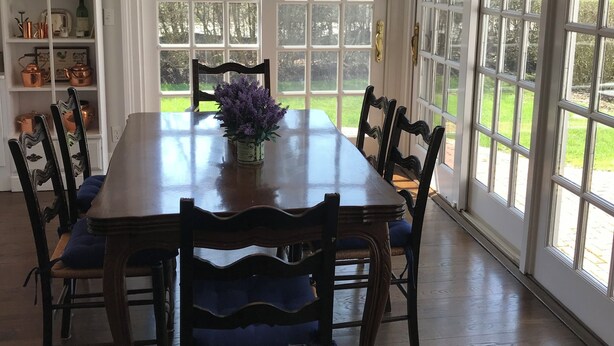 Une salle à manger avec une table en bois, des chaises, un lustre et une vue dégagée sur l’extérieur grâce à de grandes fenêtres.