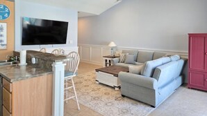 Een moderne keuken met een flatscreen televisie, een eethoek met een witte tafel en stoelen, een grijze bank en een plafondventilator.