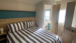 A bedroom with a bed, a wooden headboard, a nightstand, a window, and a door. - Coworth Deluxe Cabin With Garden (Carnforth)