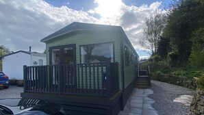 A small, single-story mobile home with a balcony, a gravel driveway, and a stone wall. - Coworth Deluxe Cabin With Garden (Carnforth)