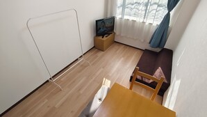 Ein Zimmer mit Holzboden, einem Tisch, einem Stuhl, einem Fernseher auf einem kleinen Schrank und einem Fenster mit Vorhängen.