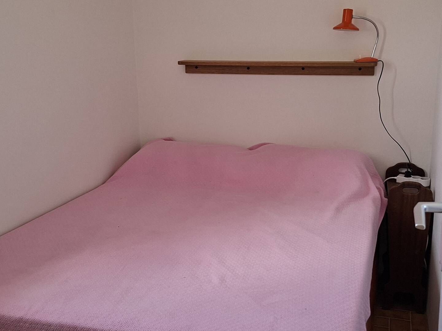 Un lit simple avec une couvre-lit rose, une étagère en bois au-dessus et une lampe rouge fixée au mur.