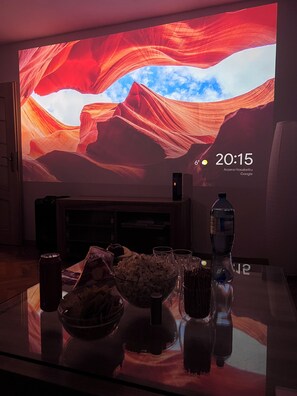 A room with a large screen displaying a scenic image, a glass table with snacks, a bottle, and a speaker. - Spacious apartment in the heart of Wroclaw – Nozownicza. 3 rooms with a loggia (Wrocław)