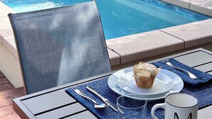 Une table placée au bord de la piscine, avec une assiette, un muffin et une tasse, entourée de palmiers et d’une clôture en bois.