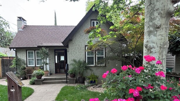 Une maison à deux étages avec un porche à l’avant, une cheminée bien visible et un jardin orné de fleurs roses.
