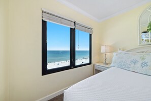 Ein Schlafzimmer mit einem Bett, einem Nachttisch, einer Lampe, einem Fenster mit Jalousien und Blick auf den Strand.