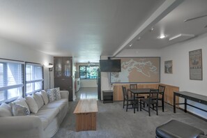 A modern living room with a sofa, coffee table, dining table, chairs, and a flat-screen TV mounted on the wall.