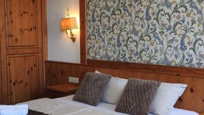 Hotellihuone, jossa on puinen sängynpääty, kuvioitu tapetti, seinään kiinnitetty lamppu ja sänky, jonka päällä on rullalle taiteltuja pyyhkeitä.
