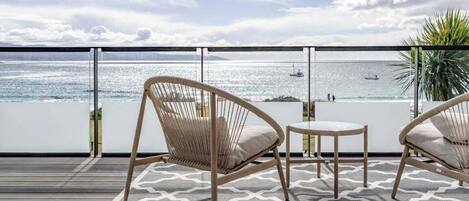 Een balkon met twee rieten stoelen en een klein tafeltje, met uitzicht op een rustige zee met een boot en land in de verte.