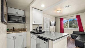 Una cocina compacta con armarios blancos, microondas, cafetera y lavavajillas. Hay una pequeña zona de estar con un sofá negro y un televisor.