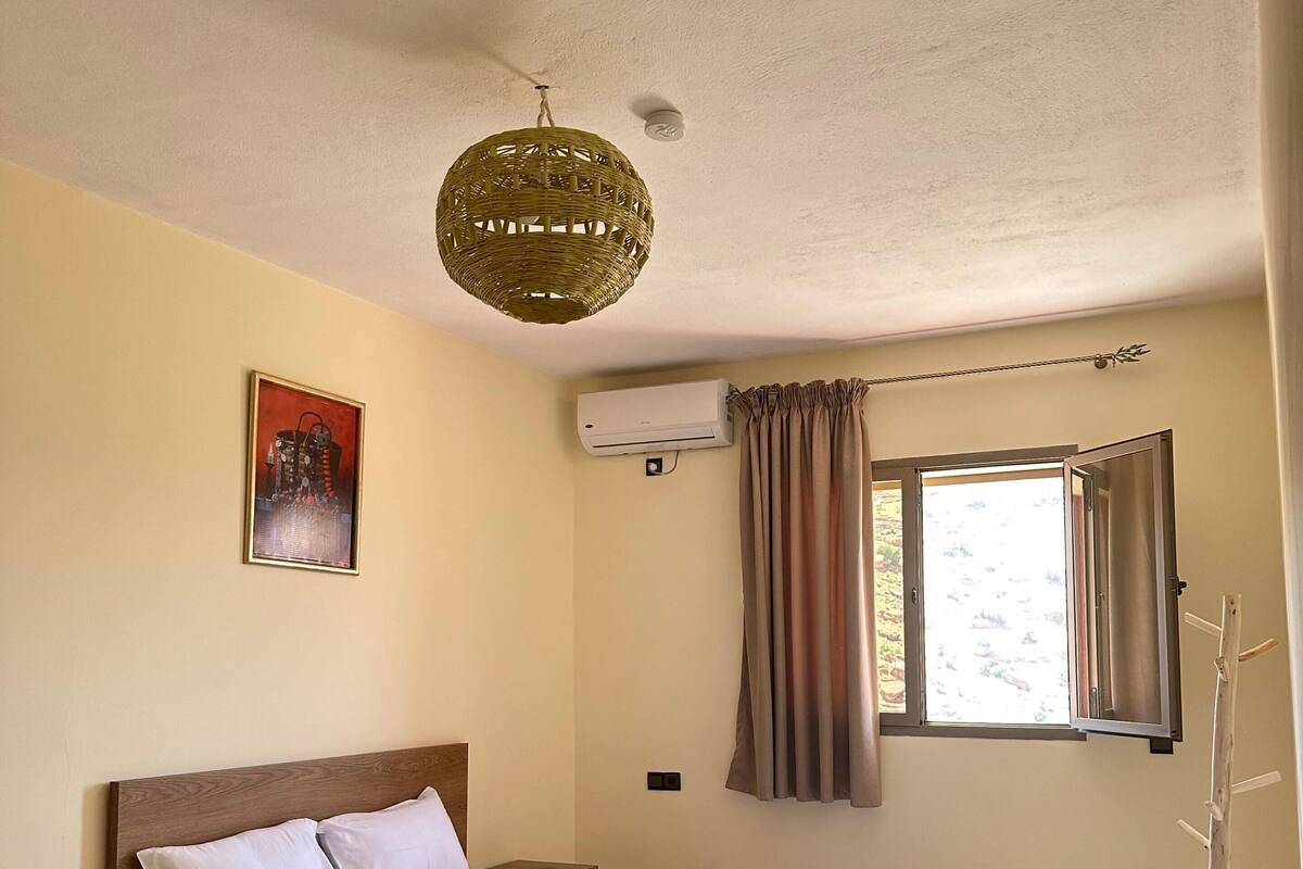 A bedroom with a bed, a nightstand, a wall-mounted air conditioner, a window with curtains, and a hanging light fixture.