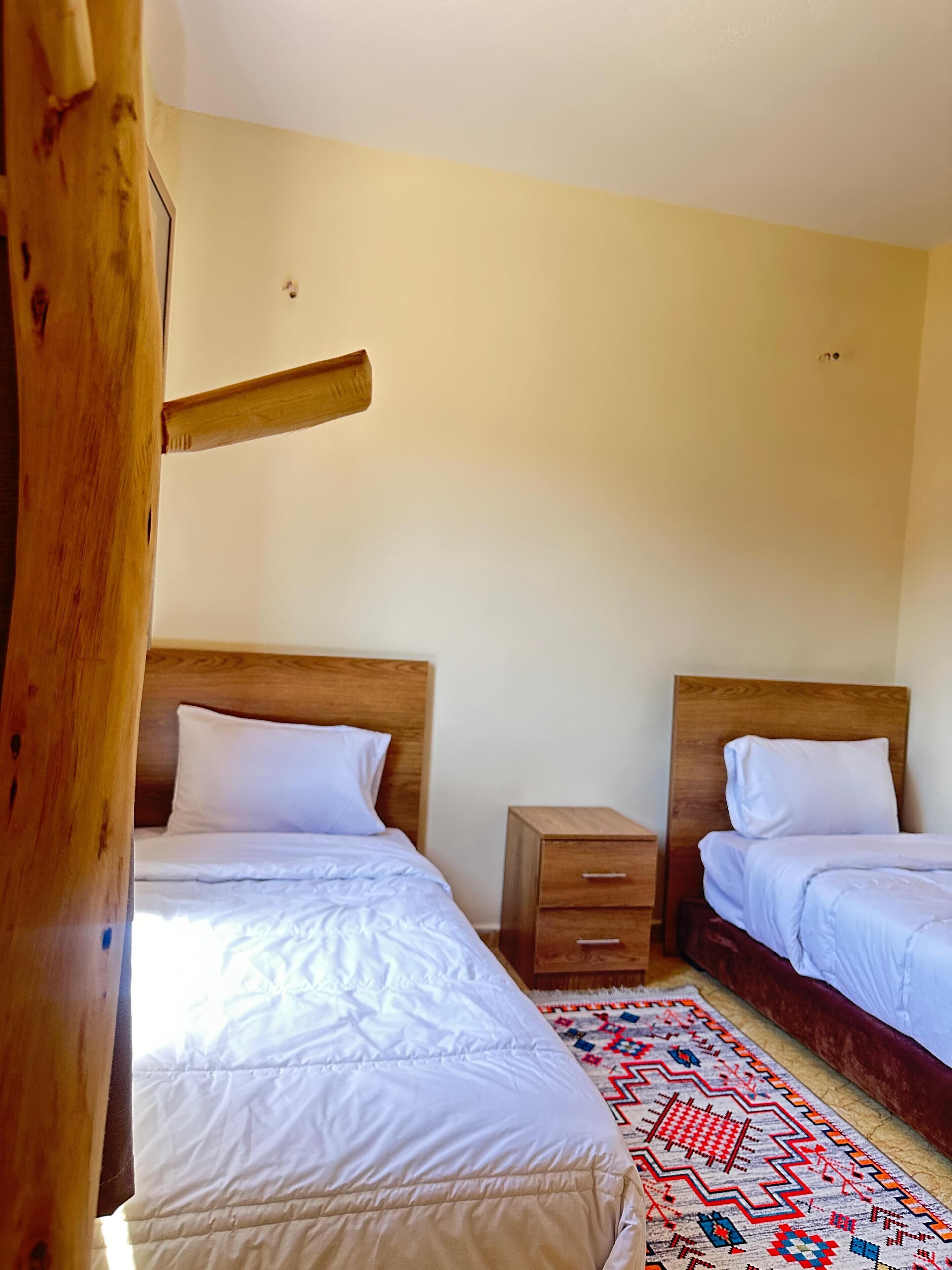 A room with two beds, wooden headboards, a small bedside table, and a patterned rug.