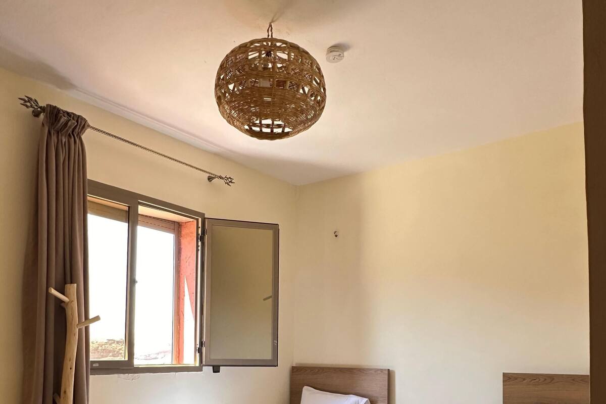 A hotel room with two beds, a wooden headboard, a small bedside table, a hanging light fixture, a window with curtains, and a wooden ladder.