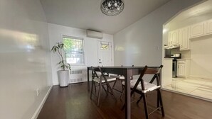 Un comedor con una mesa y sillas, una planta en maceta, una ventana con persianas y una cocina al fondo.