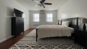 Un dormitorio con cama, ventilador de techo, cómoda, televisor y ventana con persianas.