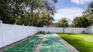 A green shuffleboard court with white markings, surrounded by a white fence and a grassy area. - Sunny Poolside Retreat | Near Beach & Dining (Clearwater)