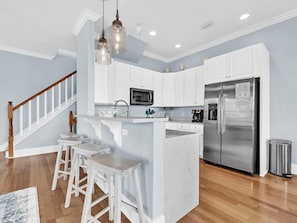A kitchen with white cabinets, a stainless steel refrigerator, a microwave, and a coffee maker. There are two white barstools and a wooden staircase. - La Valencia Beach House (Panama City Beach)