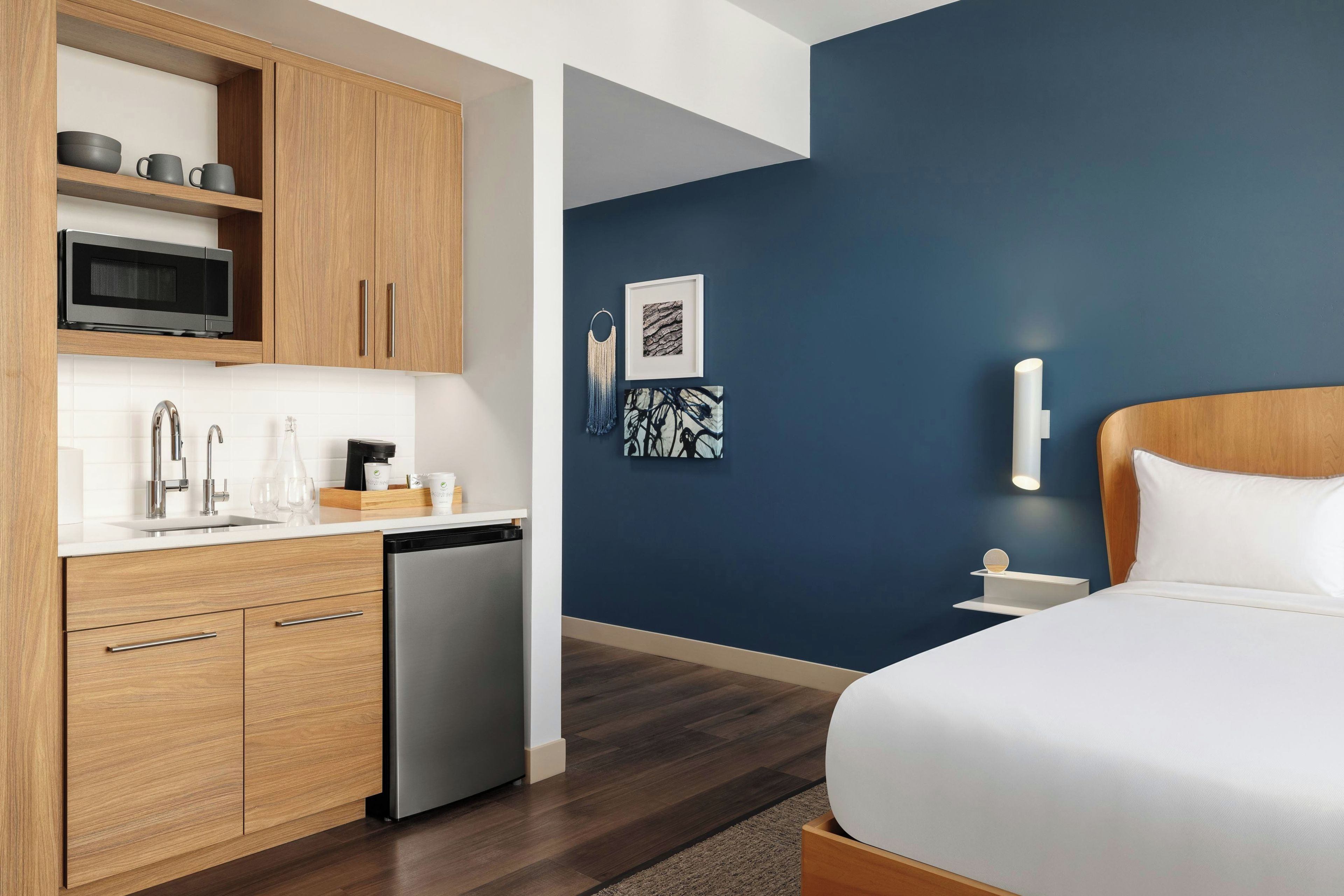 Ett modernt hotellrum med ett litet kök, en säng med vita sängkläder och konstverk på väggen.