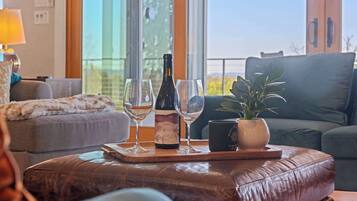 Un salon avec un canapé, une table basse sur laquelle se trouve un plateau avec une bouteille de vin et des verres, une plante en pot et une vue dégagée grâce à de grandes fenêtres.