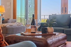 Ein Wohnzimmer mit einer Couch, einem Couchtisch mit einer Auflage, die eine Weinflasche und Gläser trägt, einer Topfpflanze und Blick durch große Fenster.