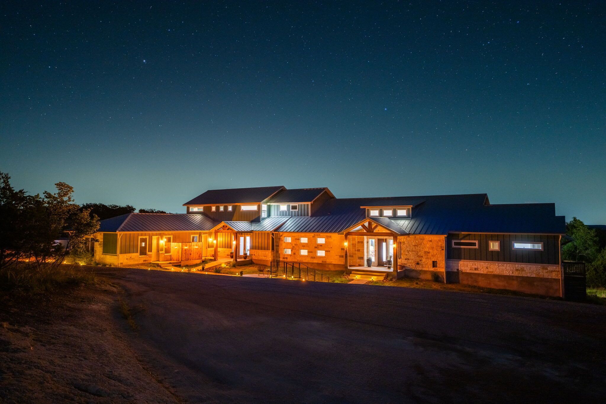 A multi-story building with illuminated windows and a well-lit entrance, surrounded by a dirt road and trees.